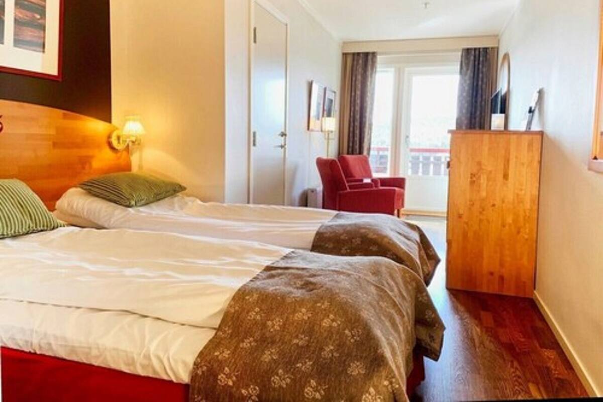 Habitación de hotel con dos camas, una silla roja, un armario de madera y un balcón con cortinas.
