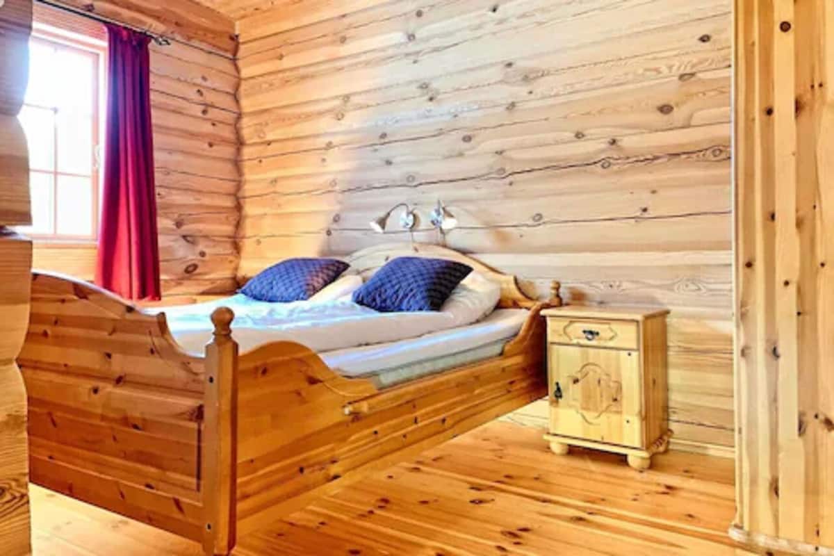 Una habitación de cabaña de madera con una cama, una mesita de noche y un ventanal con cortinas rojas.