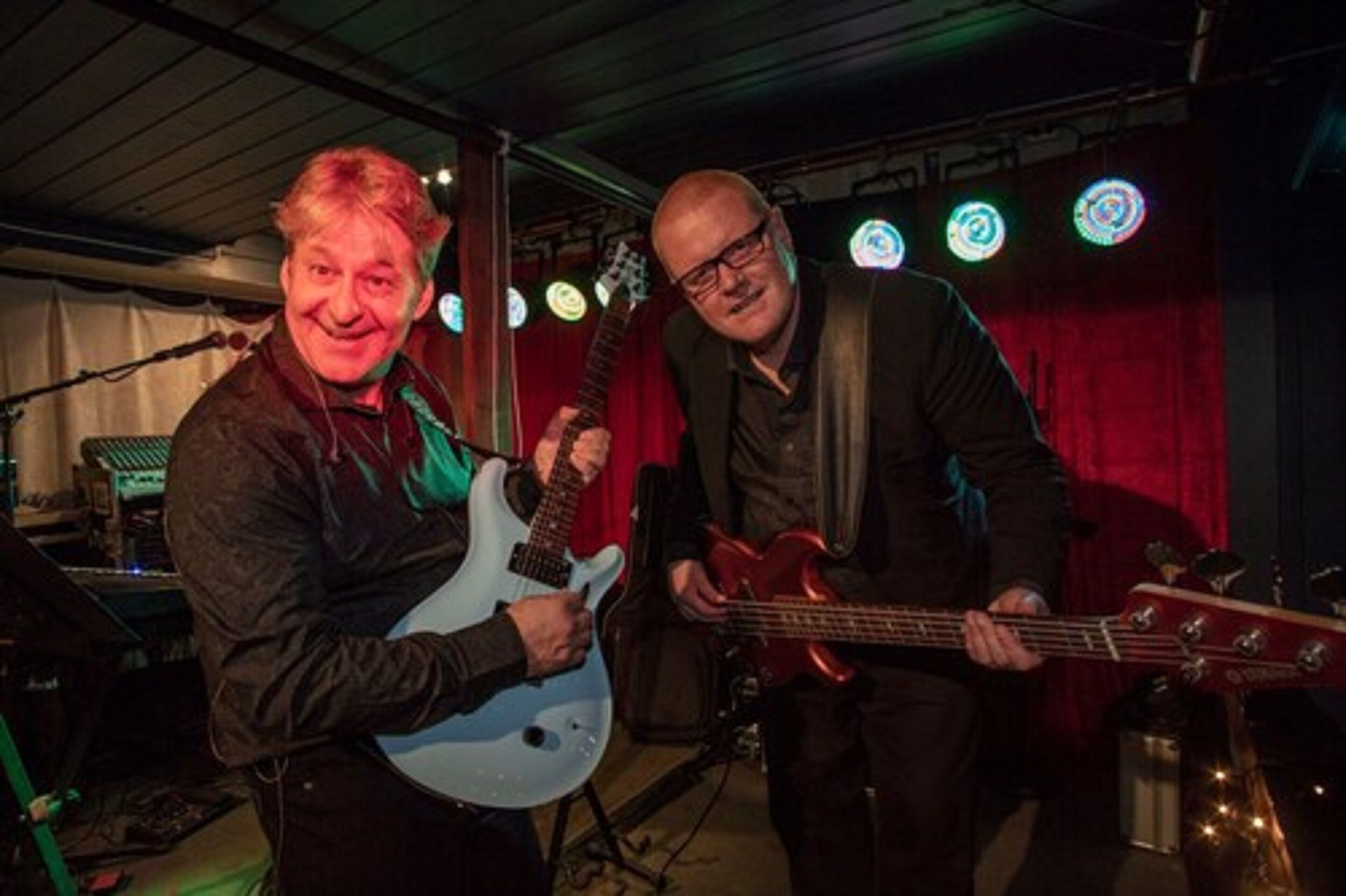 Två personer spelar elgitarr och basgitarr i ett svagt upplyst rum med en röd gardin och runda lampor.