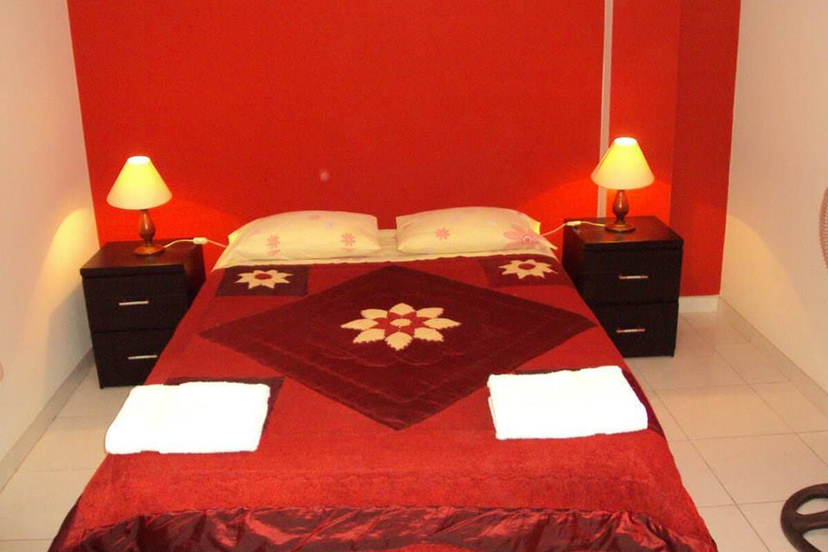 A hotel room with a red wall, a bed with a floral bedspread, two bedside tables with lamps, and a white floor.