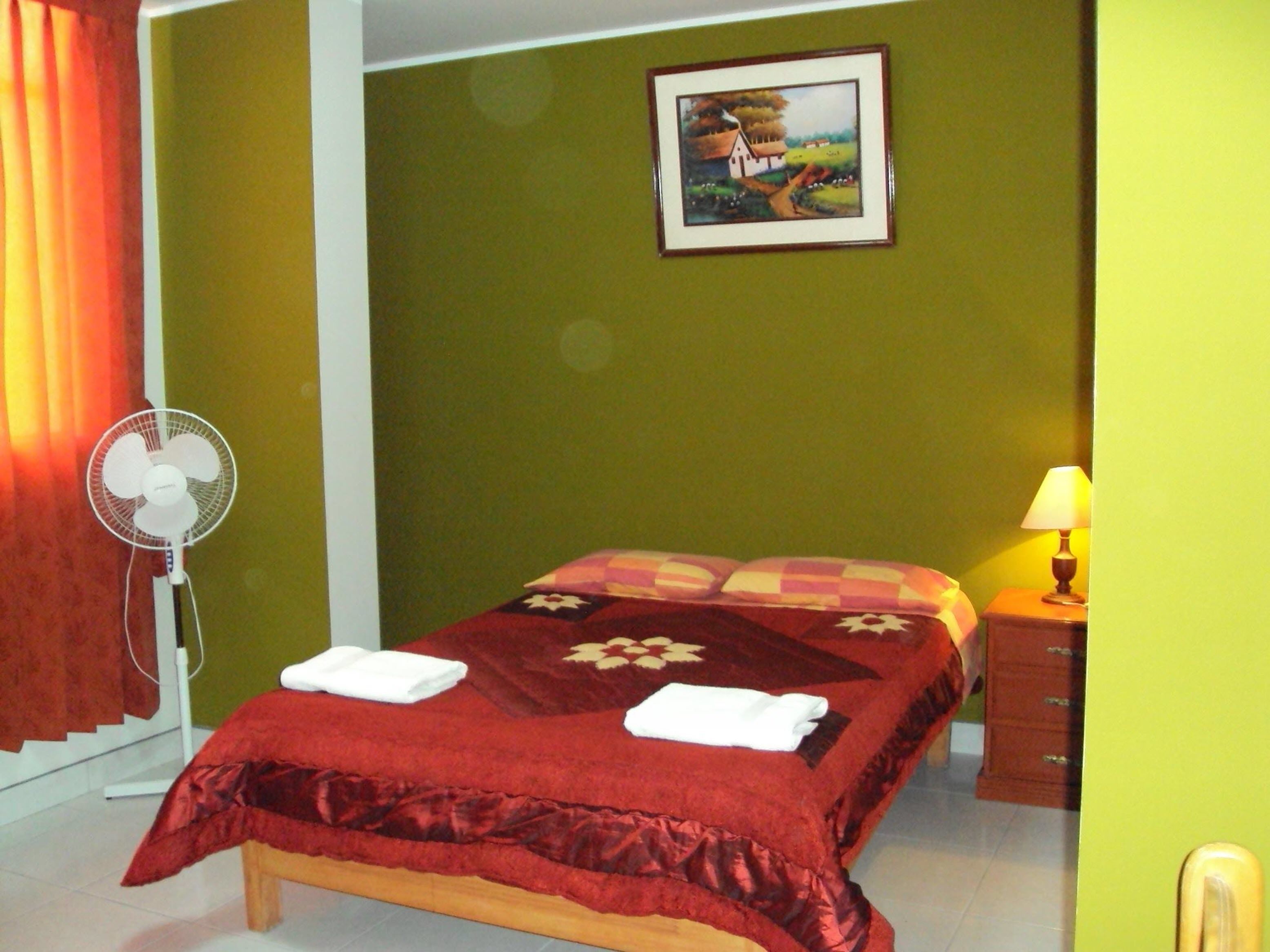 A hotel room with a bed, a bedside table with a lamp, a fan, and a painting on the wall.
