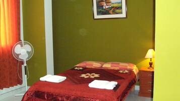 A hotel room with a bed, a bedside table with a lamp, a fan, and a painting on the wall.