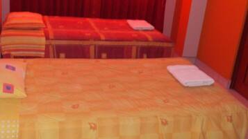 Una cama con colcha y almohadas amarillas, una mesita con una toalla doblada, y cortinas rojas al fondo.