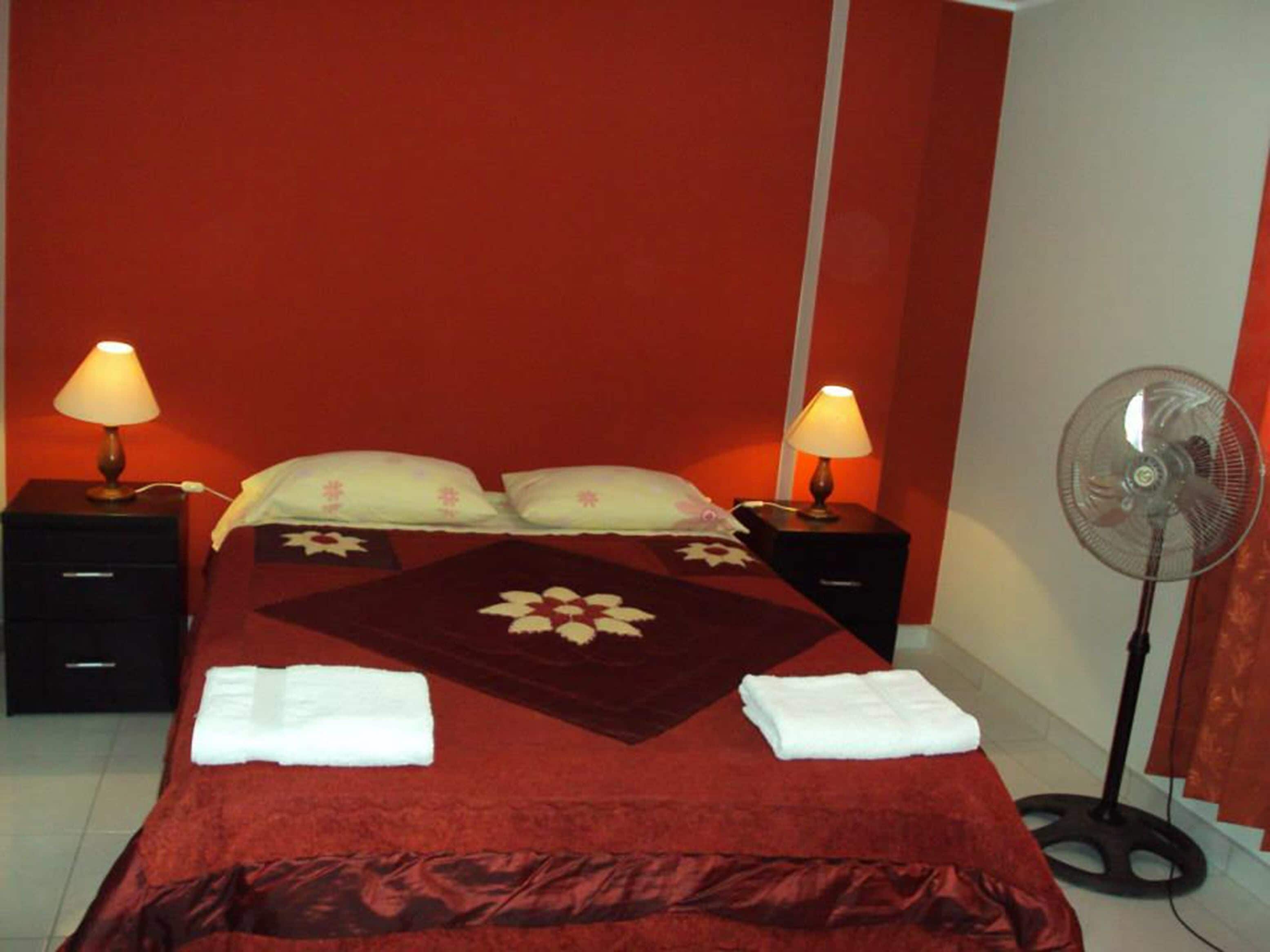 A hotel room with a large bed, two bedside tables with lamps, a standing fan, and a red wall.