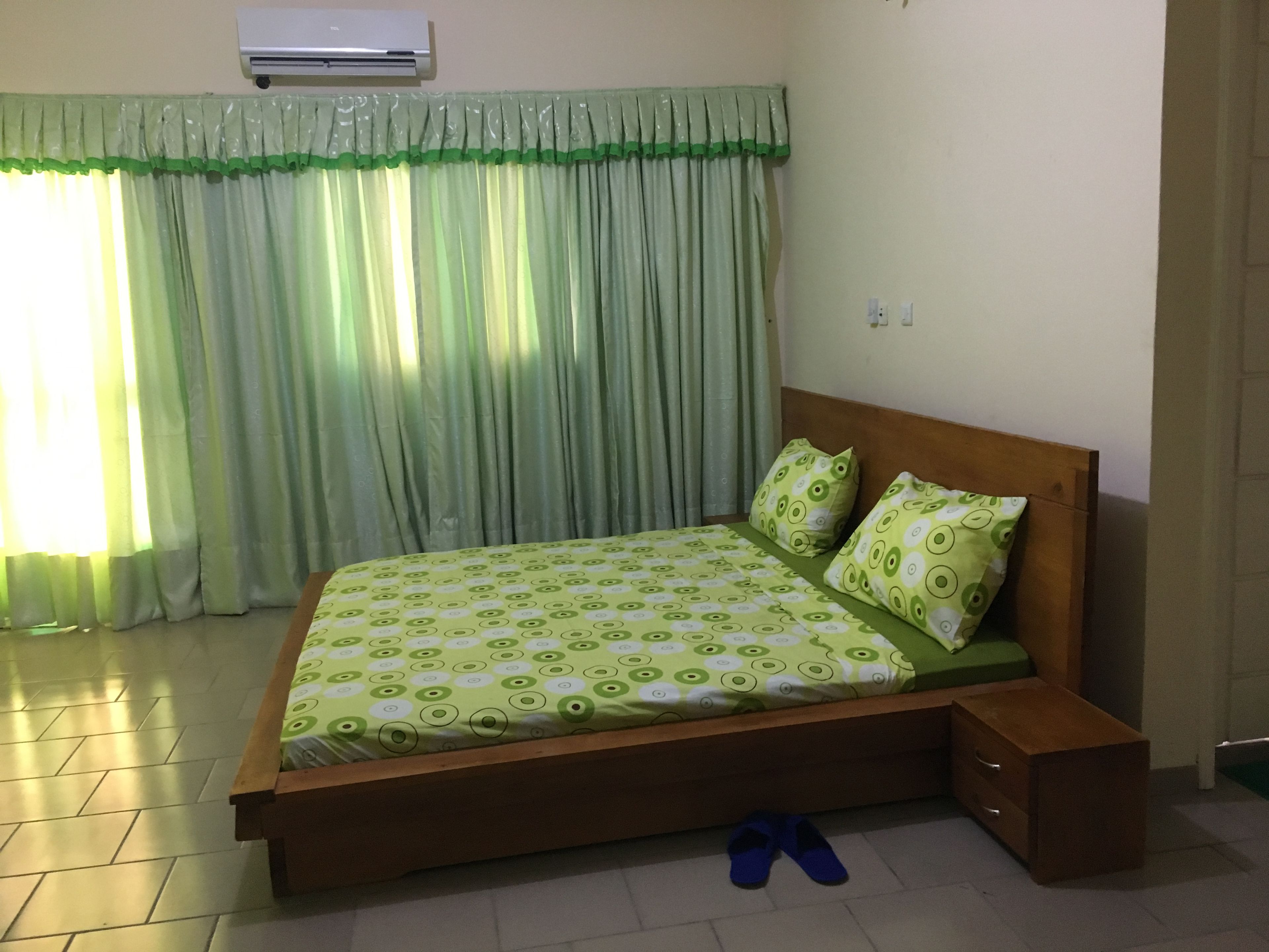 A bedroom with a bed, a nightstand, a wooden headboard, a green and white patterned bedspread, matching pillows, a pair of blue slippers on the floor, and a window with green curtains.