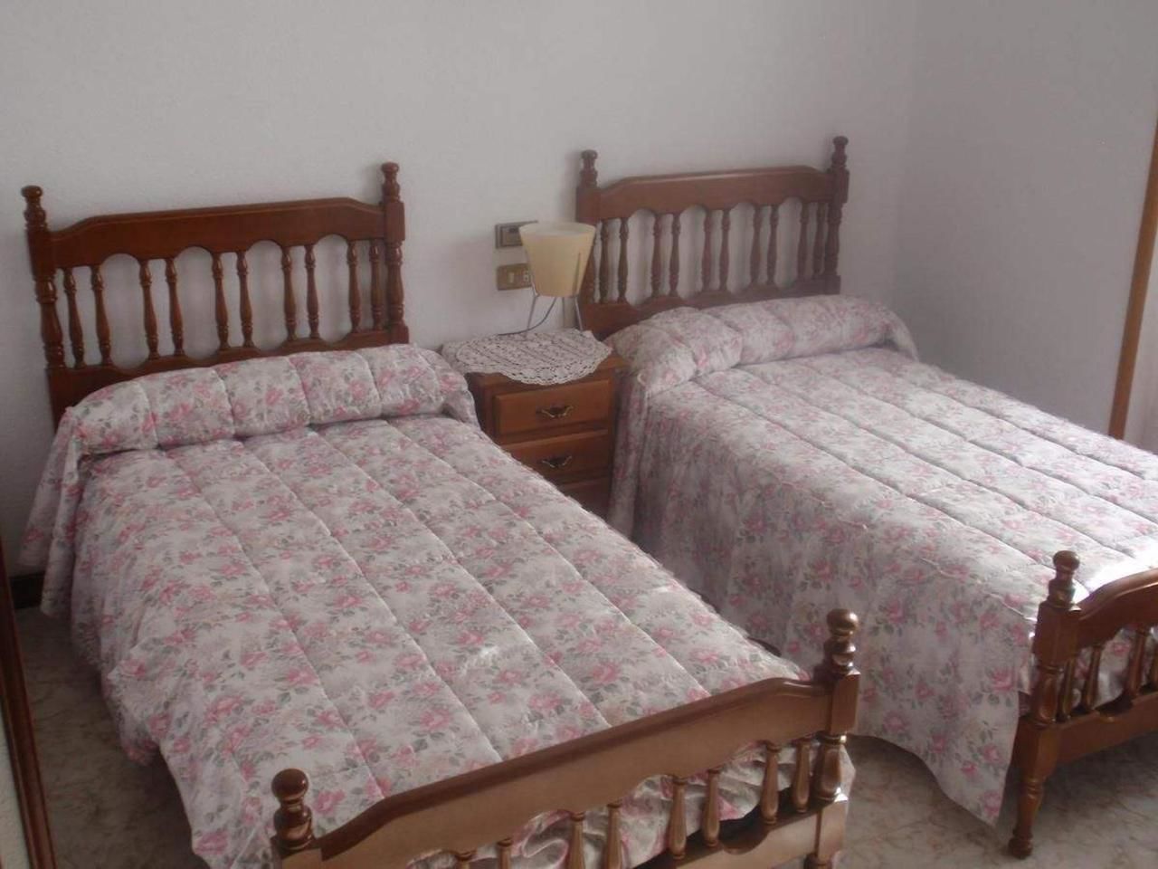 Un dormitorio con dos camas de madera, cada una con colcha estampada. Entre las camas, una mesita de noche con lámpara.