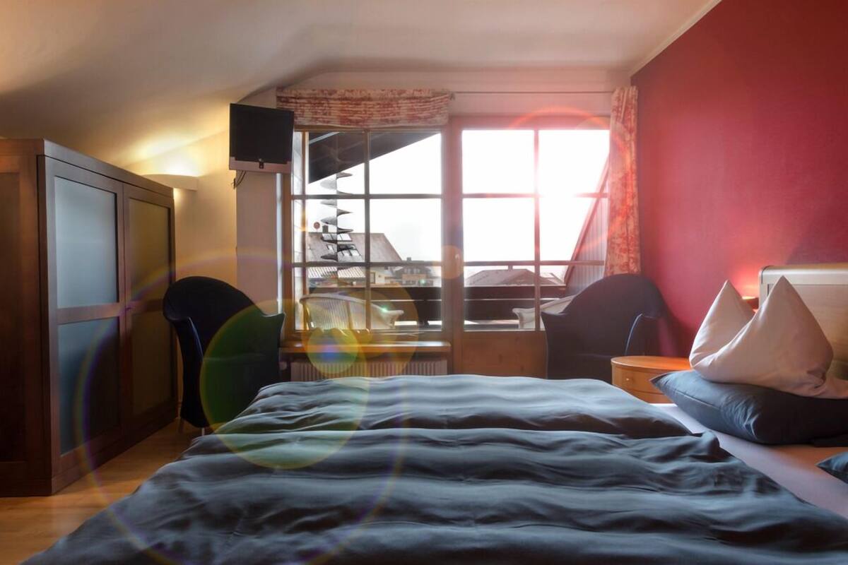 Un dormitorio con cama, escritorio, silla y ventana con vista a edificios.