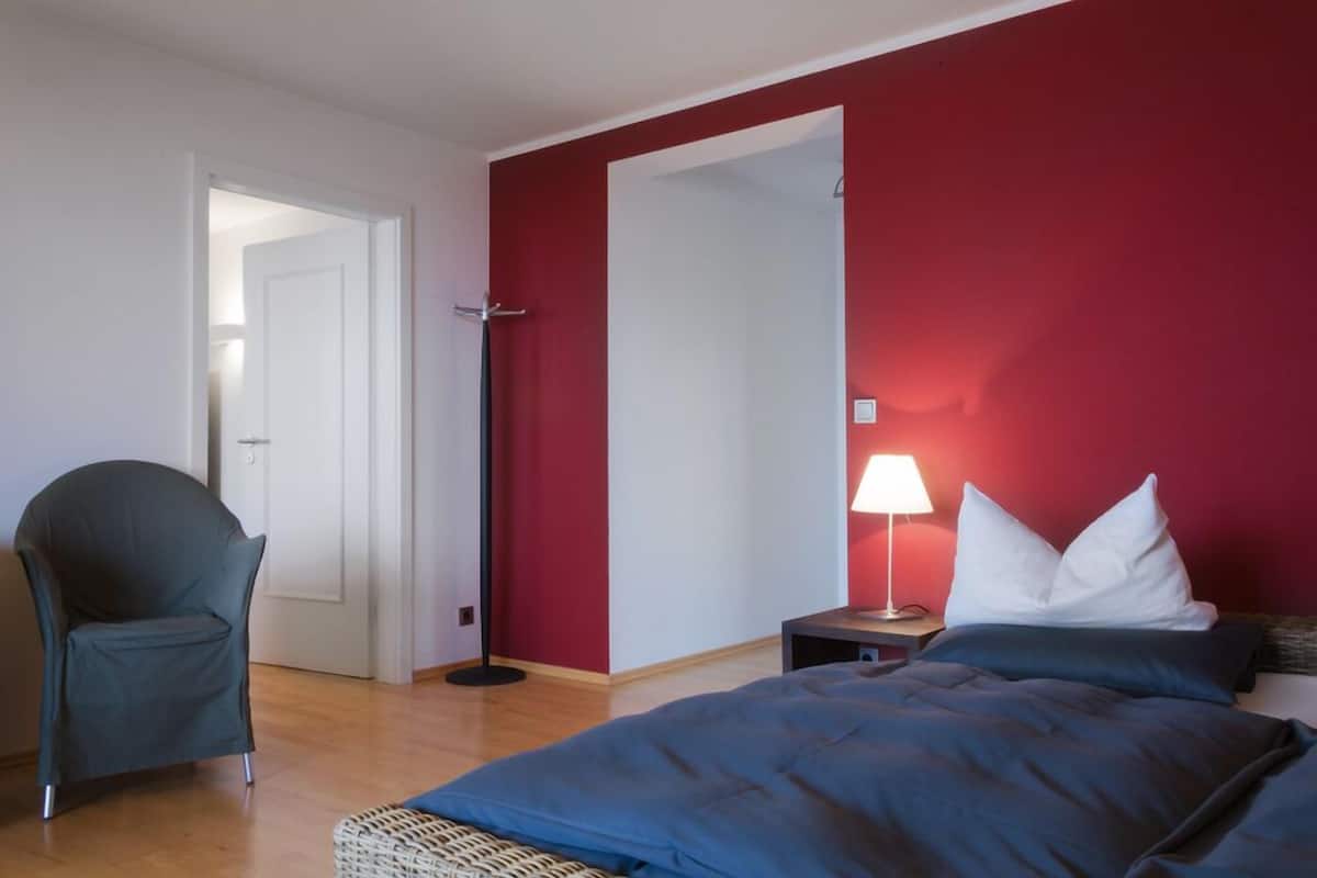 Un dormitorio con una cama, una silla, una mesita de noche con lámpara y una puerta que da a otra habitación.