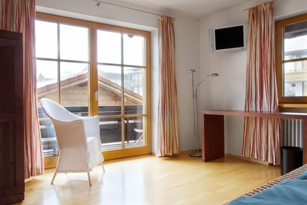 Una habitación con un gran ventanal corredera que da a un balcón, una silla blanca, un escritorio, un televisor montado en la pared y una lámpara de pie.
