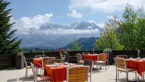 Un área de comedor al aire libre con mesas y sillas, con montañas y vegetación de fondo.