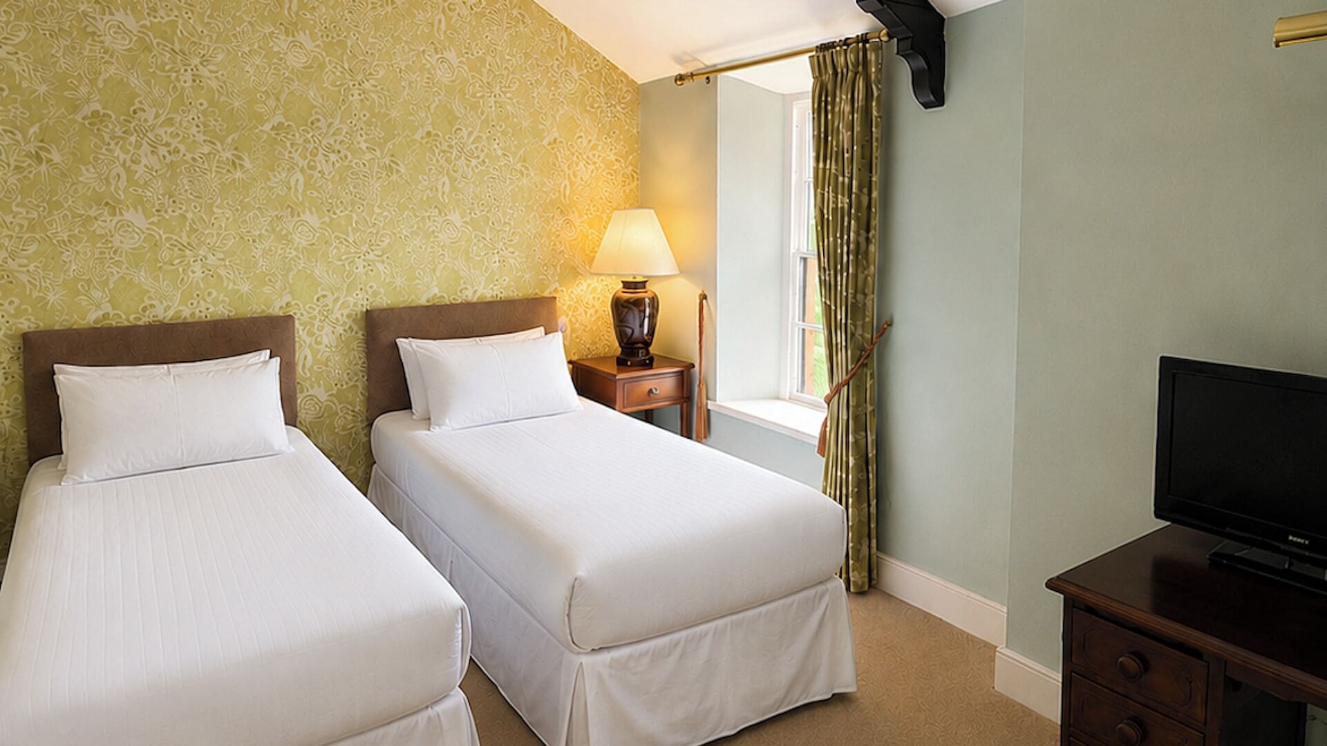 A hotel room with two single beds, a nightstand with a lamp, a TV on a wooden cabinet, and a window with curtains.