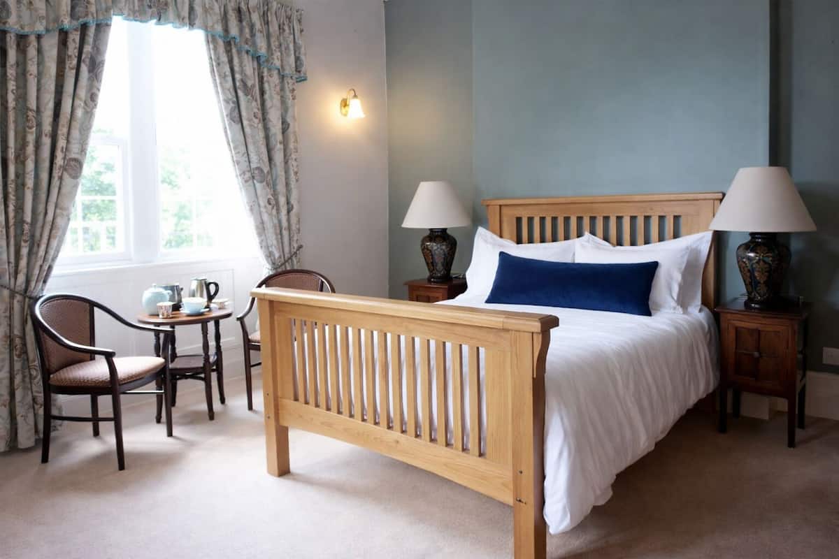 A bedroom with a wooden bed, two bedside tables with lamps, a chair, a small table, and a window with curtains.