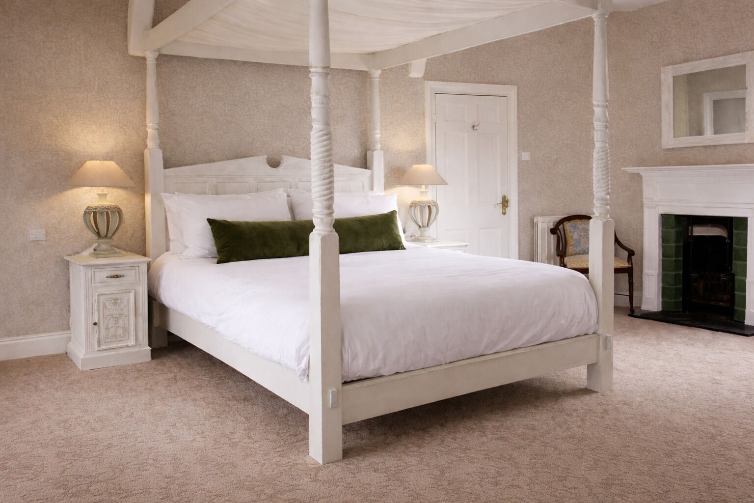 A four-poster bed with white bedding, two bedside tables with lamps, a green throw pillow, a white door, and a fireplace.