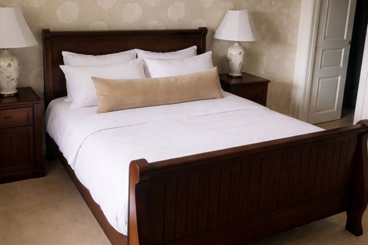 A bedroom with a wooden bed, white bedding, two bedside lamps, and a floral wallpaper pattern.