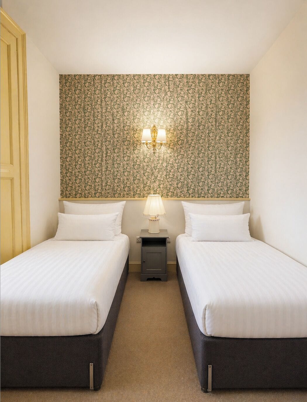 Een hotelkamer met twee bedden, behang met een patroon, een nachtkastje met een lamp en een deur aan de linkerkant.