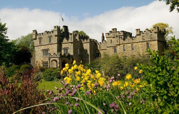 A historic stone castle with multiple towers and a flag, surrounded by lush greenery and vibrant yellow flowers.