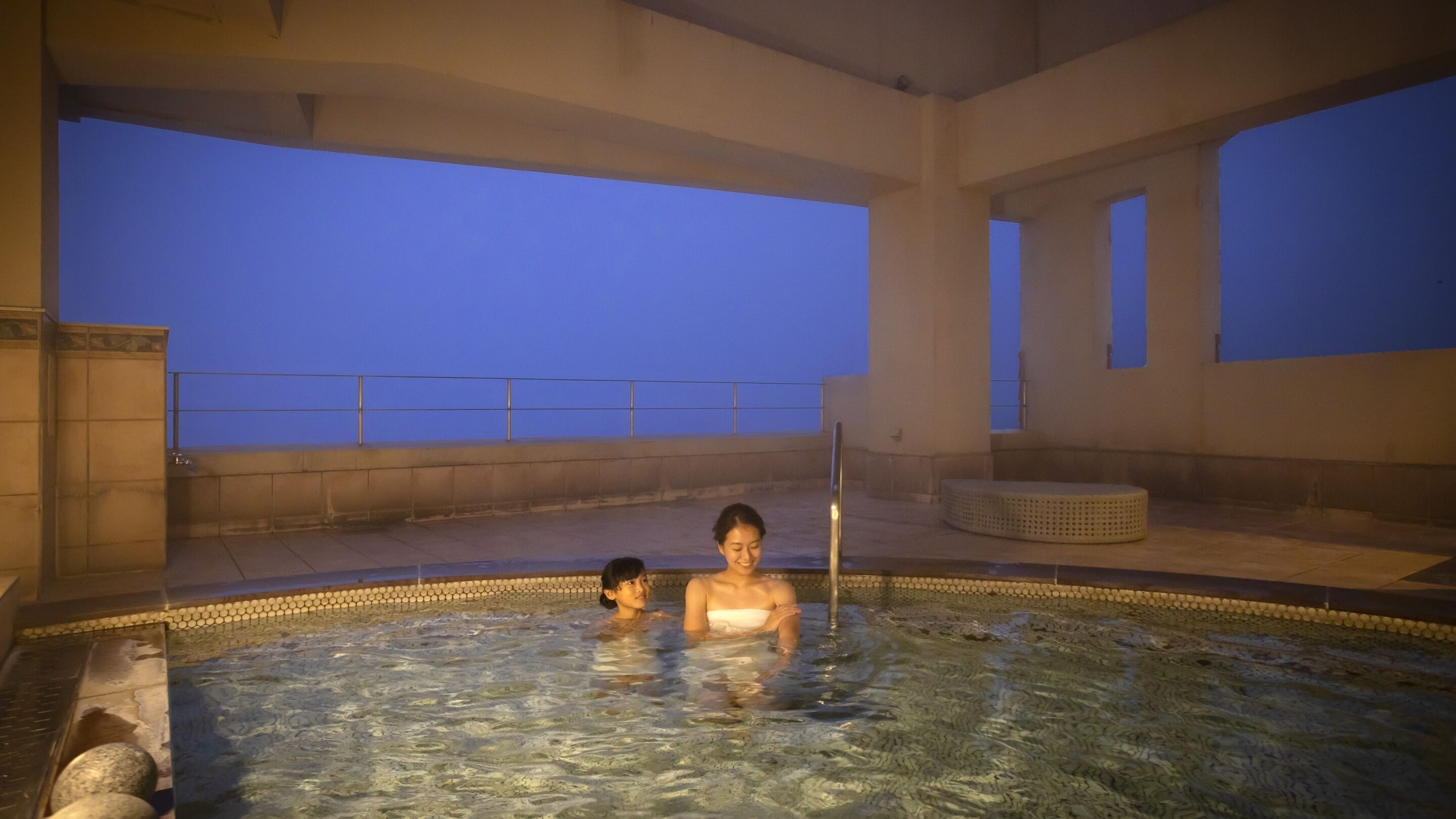 A hot tub with two people, a tiled floor, and a view of the sky.