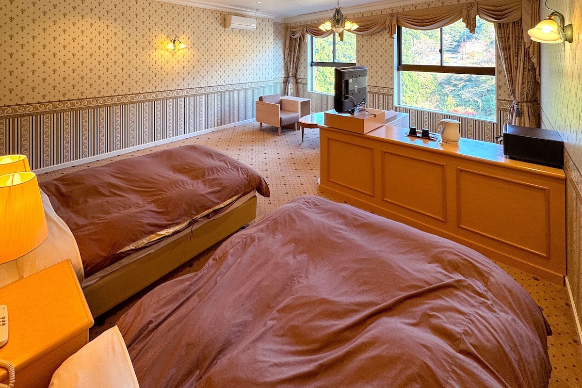 Ein Hotelzimmer mit zwei Betten, einem Schreibtisch mit Computer, einem Telefon und einem Fenster mit Vorhängen.