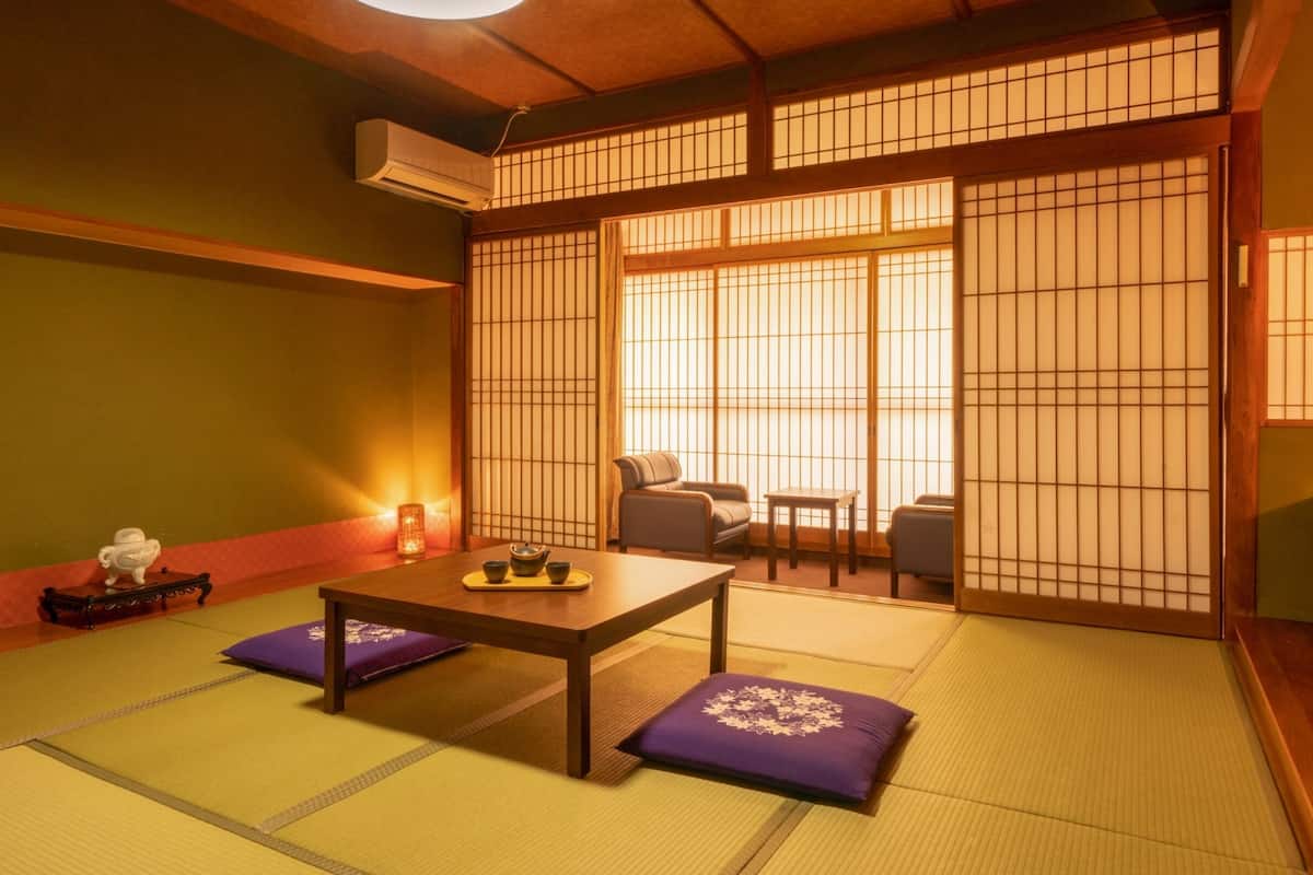 Ein traditioneller japanischer Raum mit Tatami-Bodenbelag, einem niedrigen Tisch mit Kissen, einer Schiebetür mit Shoji-Bildschirmen und einer kleinen Sitzecke mit Kissen.