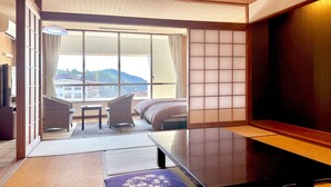 A hotel room with a sliding shoji screen, a sofa, a small table, and a view of buildings and hills. - Ooedo Onsen Monogatari Unzen Toyokan (Unzen)