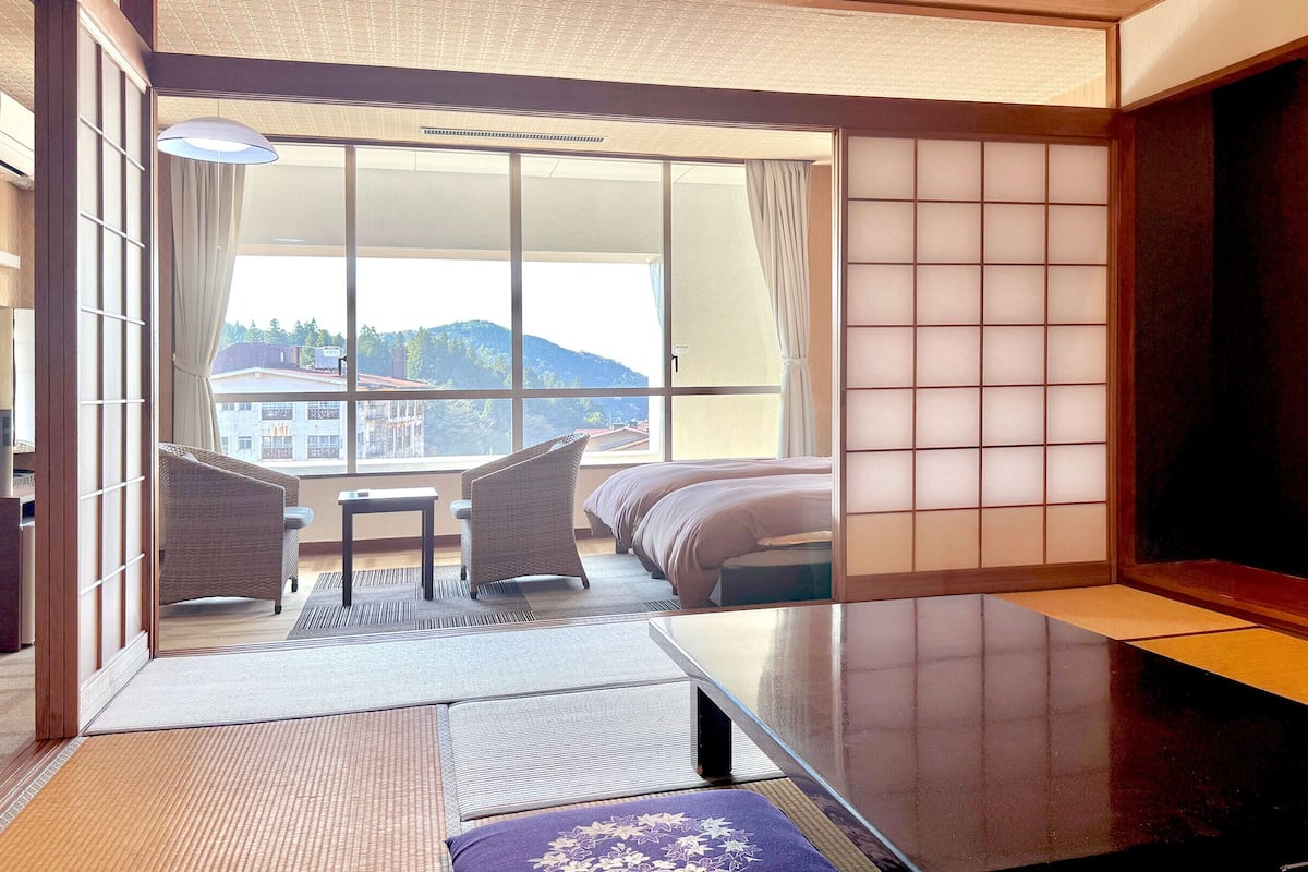 Ein Hotelzimmer mit einer verschiebbaren Shoji-Schirmwand, einer Couch, einem kleinen Tisch und Blick auf Gebäude und Hügel.