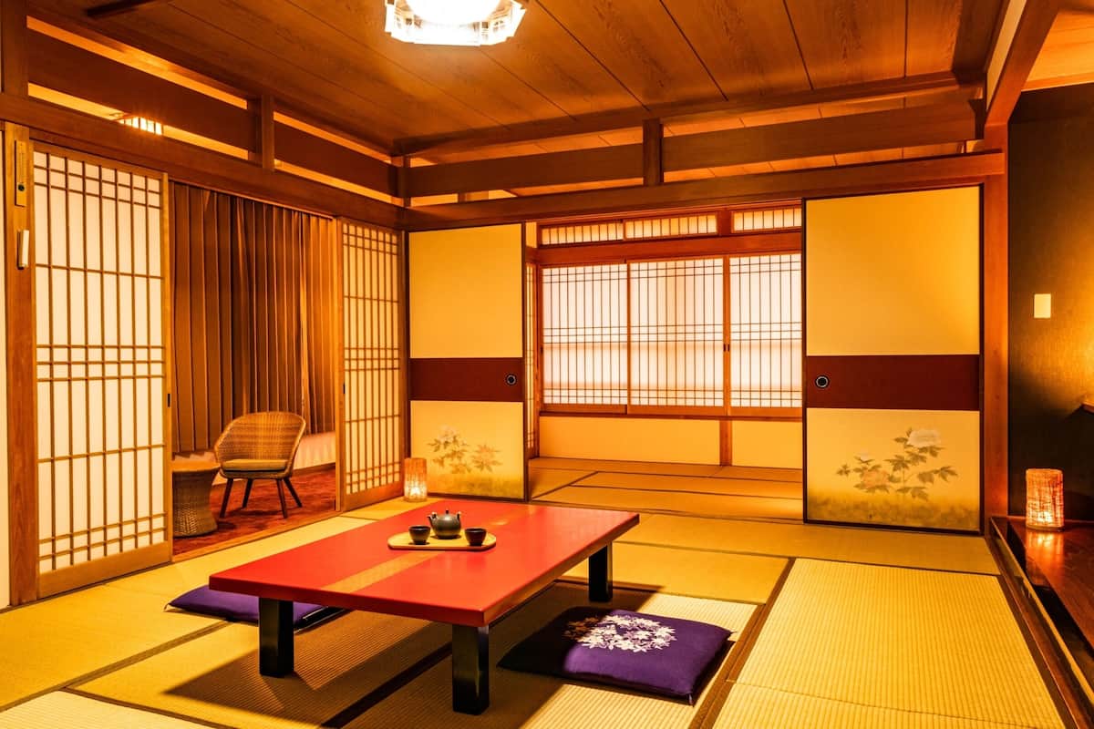 Ein traditioneller japanischer Raum mit Tatami-Böden, einem niedrigen Tisch, einem Stuhl, Schiebetüren und Blick durch einen Shoji-Bildschirm.