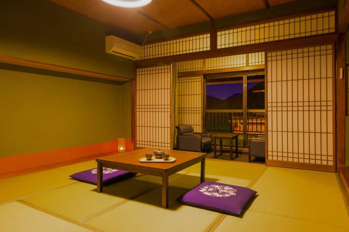 Ein traditioneller japanischer Raum mit Tatami-Böden, einem niedrigen Tisch mit Kissen, einer Schiebetür und Blick ins Freie.