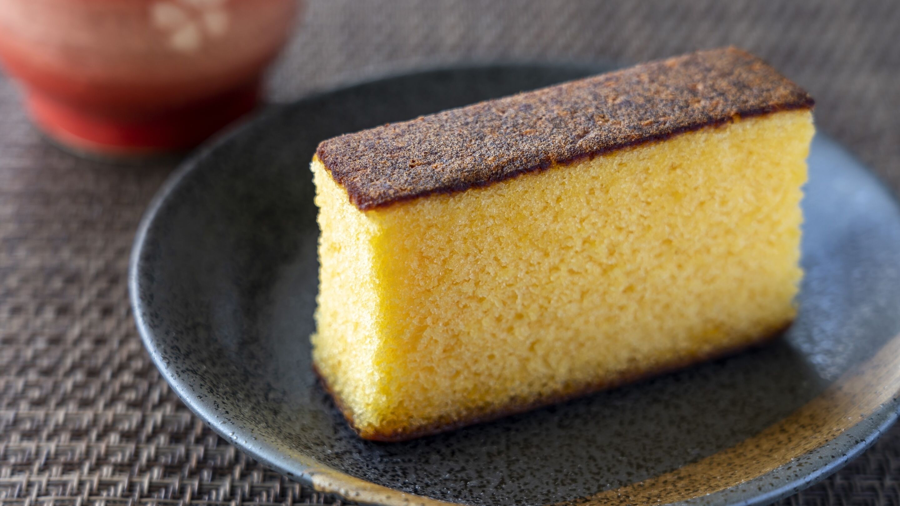 A slice of yellow cake on a dark plate.