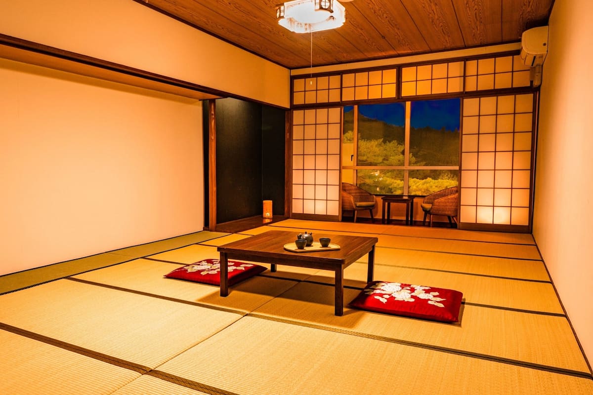 Ein Raum mit Tatami-Matten, einem niedrigen Tisch, Kissen, einer Schiebetür und Blick ins Freie.