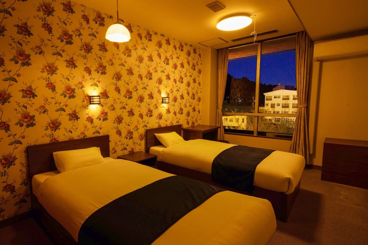 Ein Hotelzimmer mit zwei Betten, blumigem Wandpapier, Nachttischlampen, einem Fenster mit Blick auf Gebäude und einer Deckenleuchte.