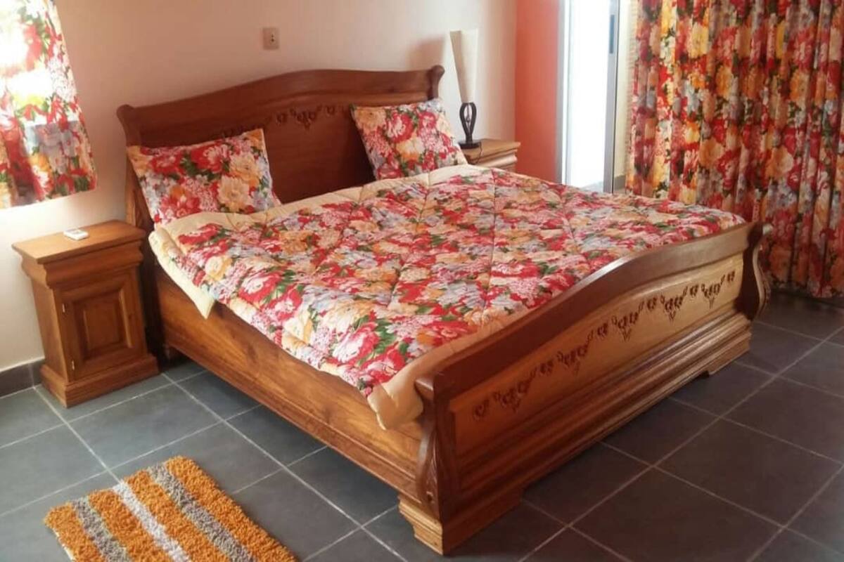 A wooden bed with floral bedding, a nightstand, and a striped rug in a room with tiled flooring.