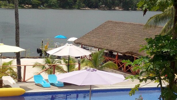 Zona junto a la piscina con sillones de descanso, sombrillas y una estructura de techo de paja.