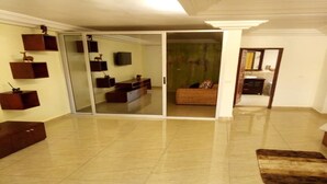 Amplia sala de estar con puerta corrediza de vidrio que da a un dormitorio. Hay un televisor de pantalla plana montado en la pared, un mueble de madera y un tapete de cuero marrón en el suelo.