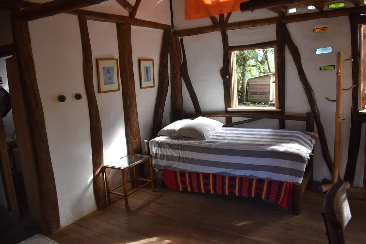 Una habitación rústica con vigas de madera a la vista, una cama con ropa de cama a rayas, una mesita y una ventana con vistas a la vegetación.
