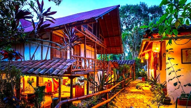 Ein Holzhaus mit Balkon, umgeben von tropischen Pflanzen und einem Kopfsteinpflasterweg.