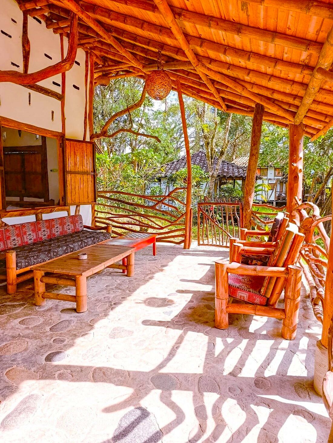 Un área de estar al aire libre con muebles de madera y suelo de piedra, rodeada por una pérgola de madera y abundante vegetación.