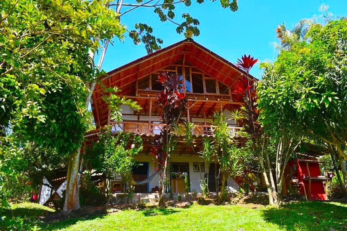Un edificio de dos plantas con tejado rojo y balcones de madera, rodeado de abundante vegetación y árboles.