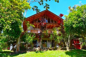 A two-story building with a red roof and wooden balconies, surrounded by lush greenery and trees.