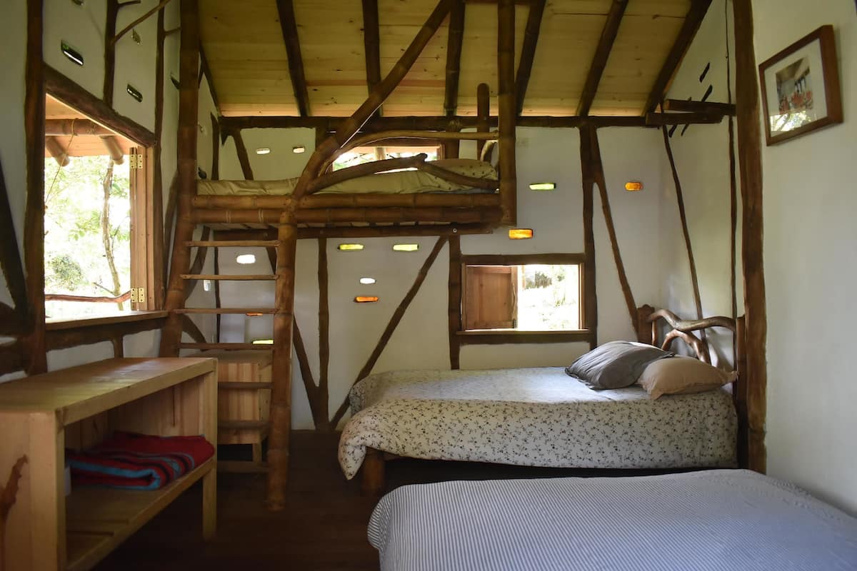 Una habitación rústica con cama alta, una escalera, un estante de madera y una ventana.