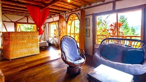 Ein Zimmer mit Holzboden, Korbmöbeln, großen Fenstern und Blick ins Grüne.