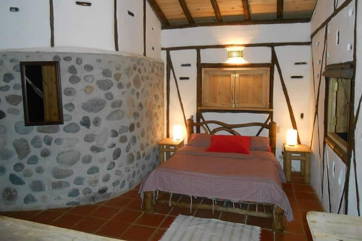 Un dormitorio rústico con pared de piedra, vigas de madera, una cama con almohada roja y un mueble de madera sobre la cama.