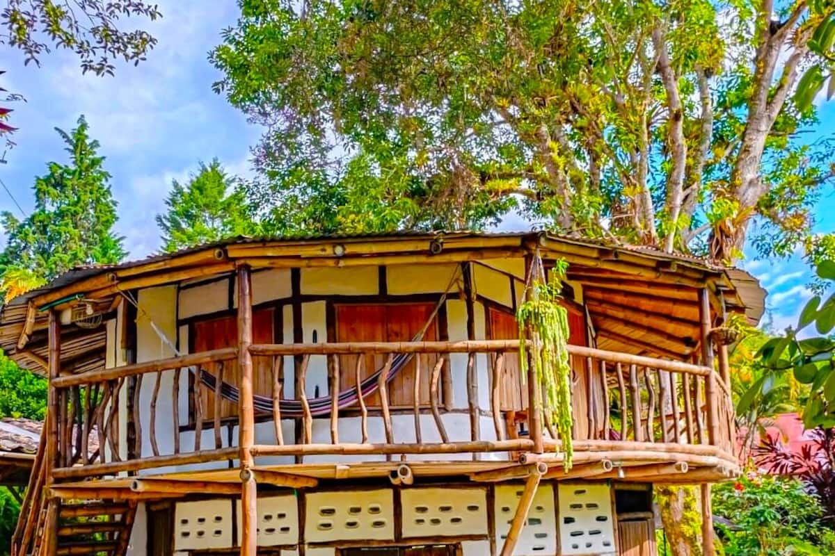 Una casa de madera de dos pisos con un balcón, rodeada de abundante vegetación.