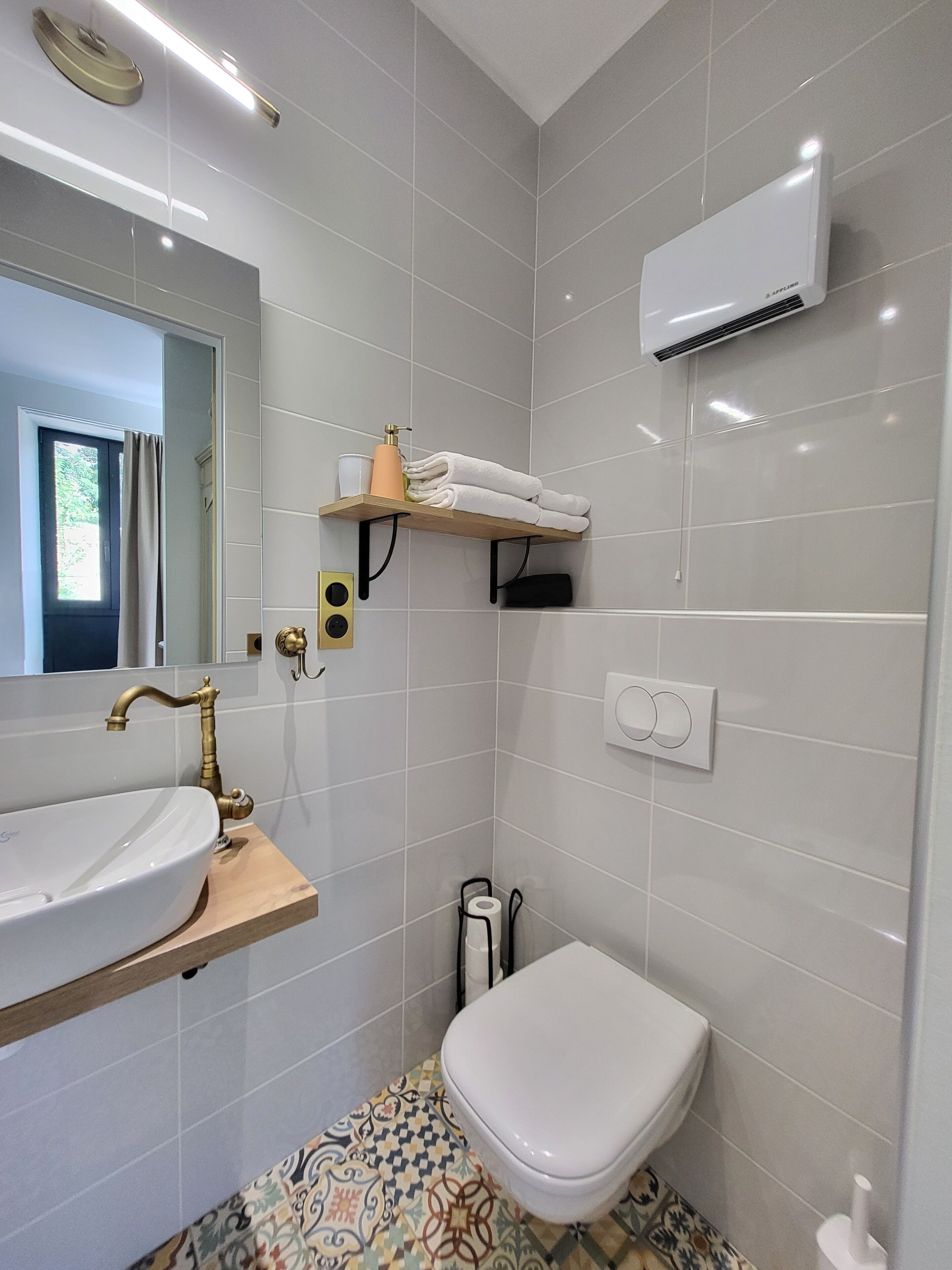 Un baño moderno con un lavabo blanco, grifo dorado y un inodoro. Hay un estante con toallas y una botella de jabón.