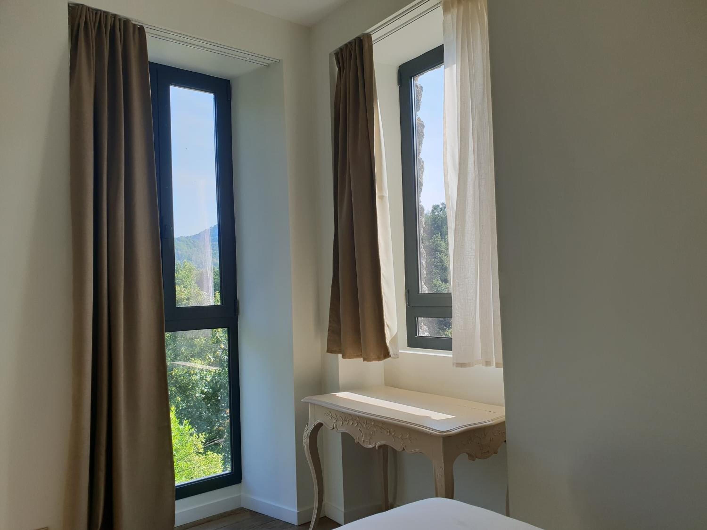 A room with two windows, one with brown curtains and another with white curtains. There is a small white table with a curved design next to the windows.