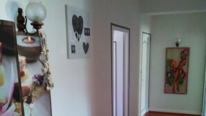 Un pasillo con suelo de madera, un banco con libros, un estante en la pared con objetos decorativos y una puerta al final.