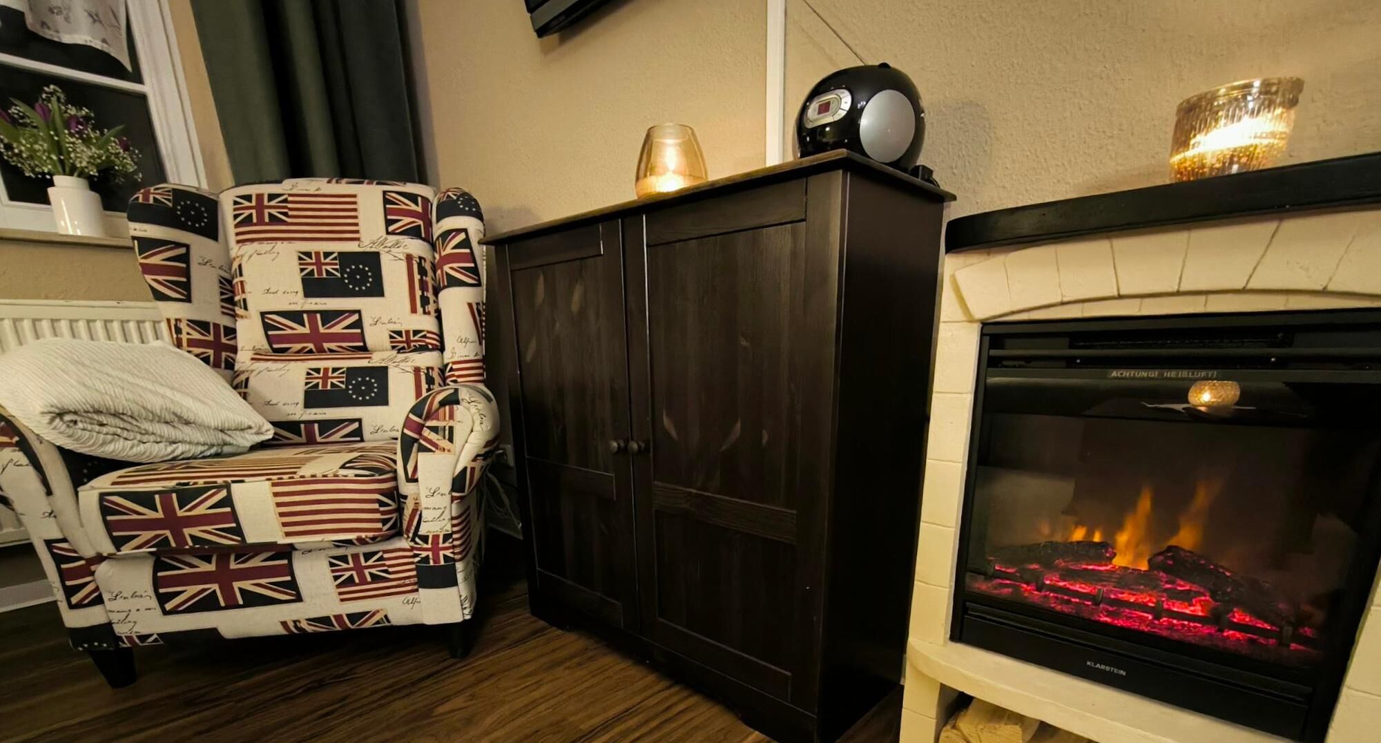 Een gezellige woonkamer met een gestreept fauteuil, een donkere houten kast en een open haard met vlammen.