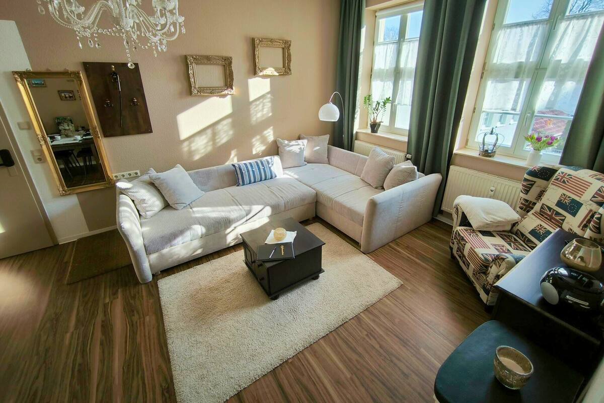 Ein modernes Wohnzimmer mit einer Eckcouch, einem Couchtisch und einem gemusterten Sessel. Es gibt eine Chandelüster, ein Fenster mit Vorhängen und einen Spiegel an der Wand.
