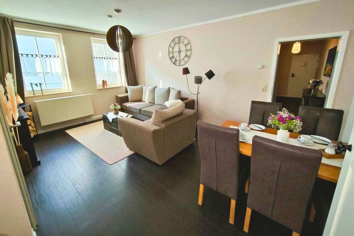 Ein modernes Wohnzimmer mit einer Couch, einem Esstisch mit Stühlen, einer Uhr an der Wand und einem Fenster mit Vorhängen.