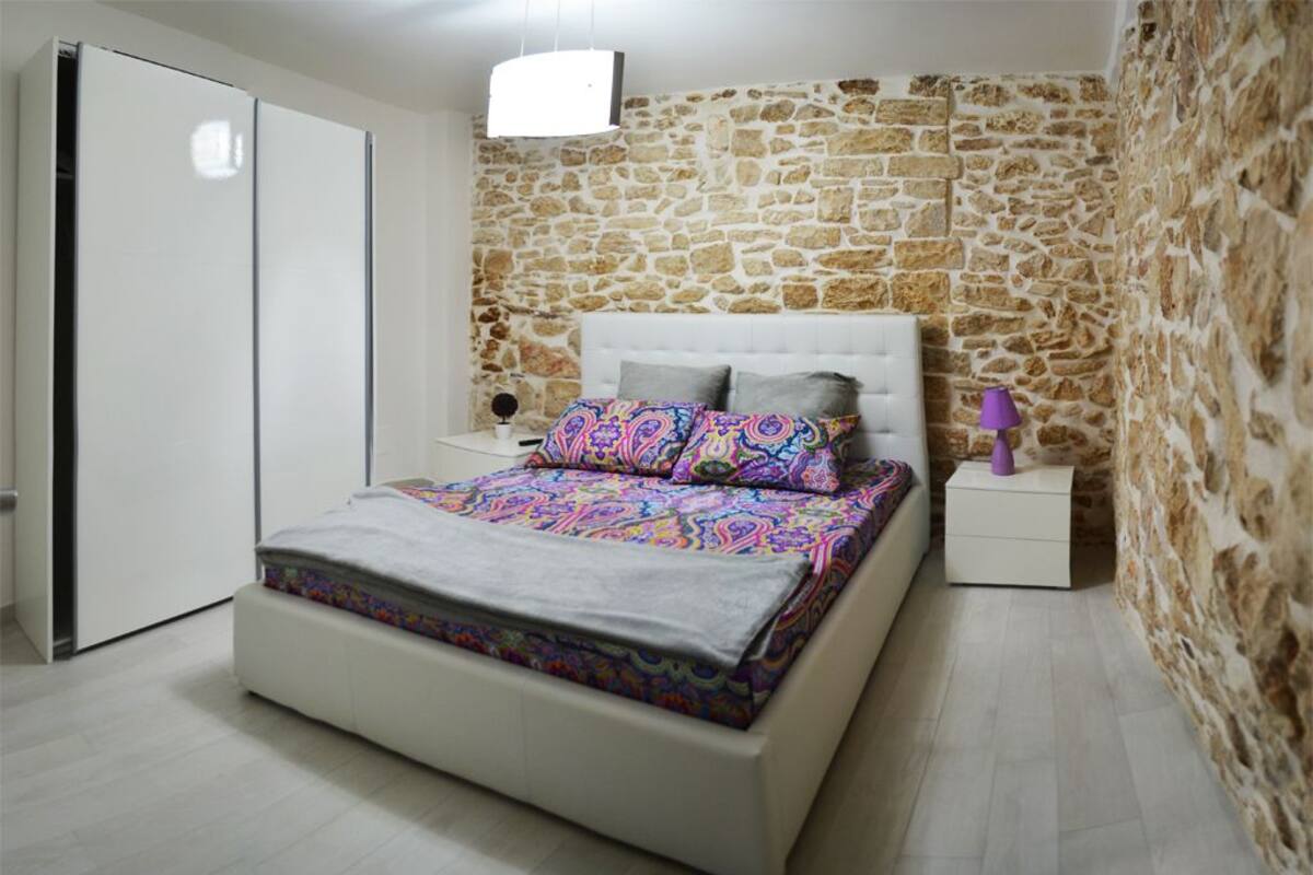 Un dormitorio con pared de piedra, cama blanca y mesita de noche con lámpara.