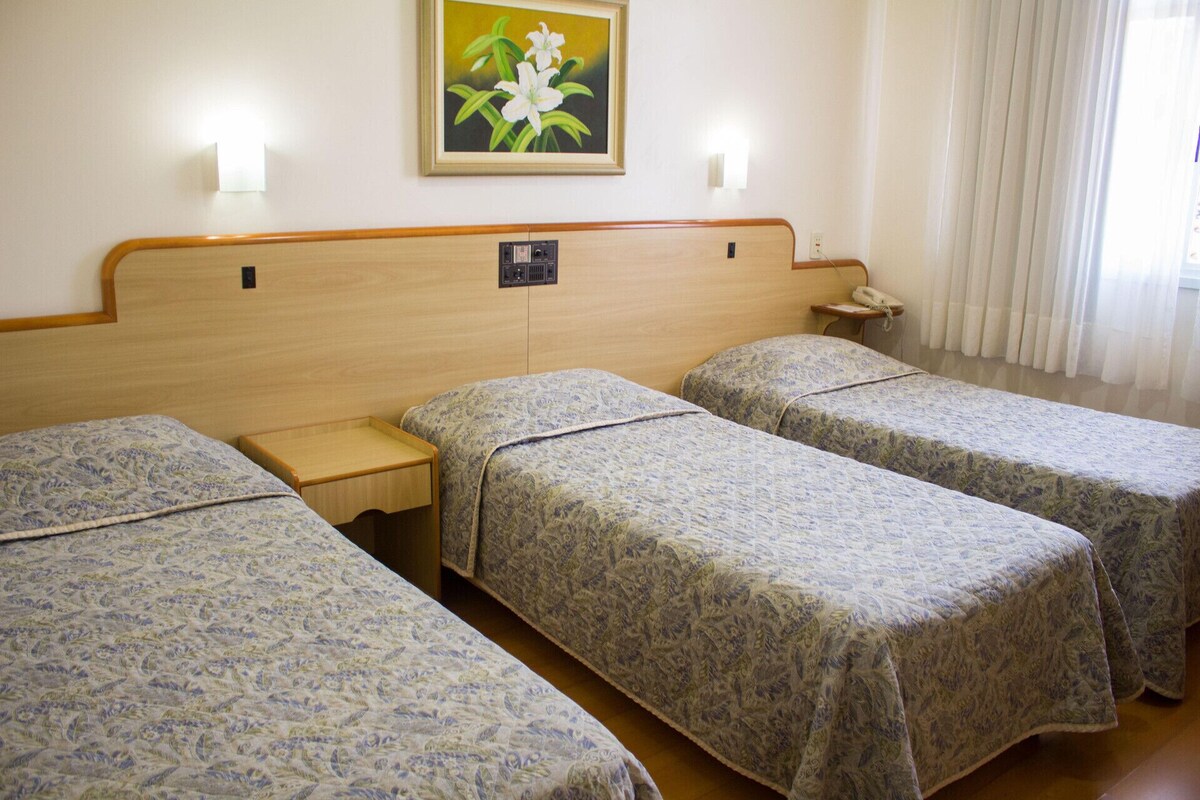 Een hotelkamer met drie eenpersoonsbedden, een nachtkastje met een telefoon, een schilderij aan de muur en een raam met gordijnen.